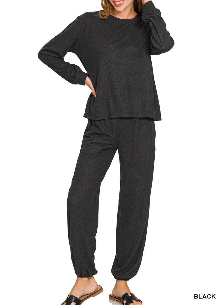 A woman modeling a black two-piece loungewear set consisting of a long-sleeve crewneck top and matching jogger pants, shown against a plain white background.