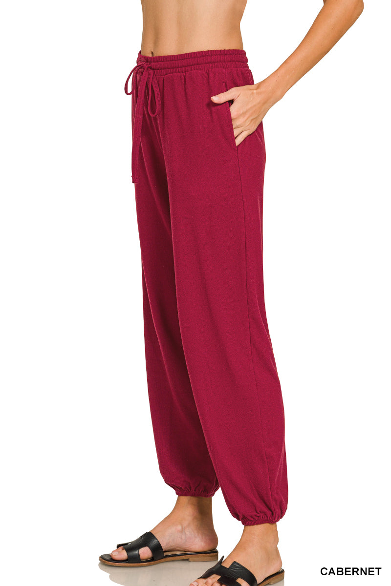 A person wearing loose harem-style pants in a deep cabernet red color, shown from the waist down against a white background. The pants have an elastic drawstring waist, side pockets, and gathered cuffs at the ankles.