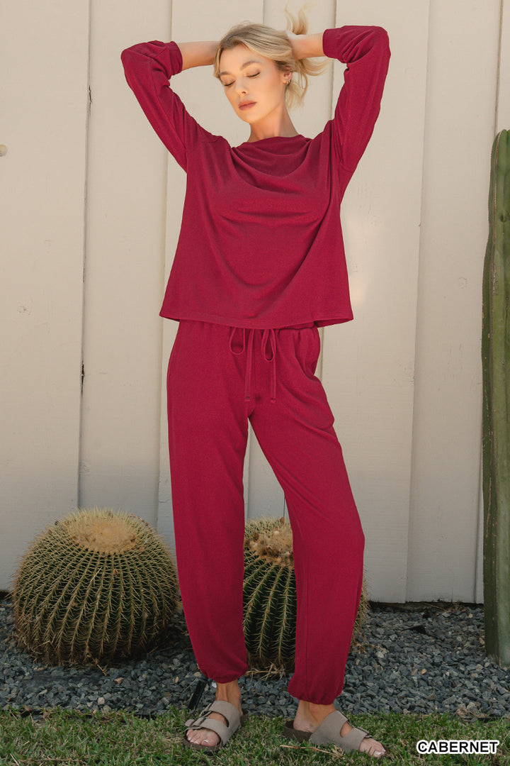 A woman modeling a matching two-piece lounge set in a deep burgundy or maroon color, consisting of a long-sleeve cropped top and drawstring pants, standing outdoors in front of a white wooden wall with cacti.