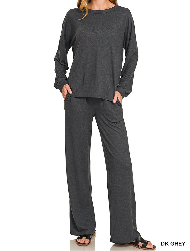 A woman modeling a dark grey two-piece loungewear set consisting of a long-sleeve top and matching wide-leg pants, shown against a plain white background with &
