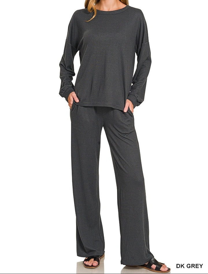 A woman modeling a dark grey two-piece loungewear set consisting of a long-sleeve top and matching wide-leg pants, shown against a plain white background with &