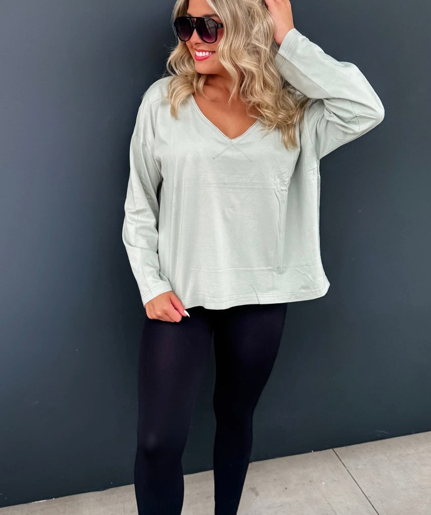 Woman wearing a light gray long-sleeve top and black leggings against a dark gray wall.