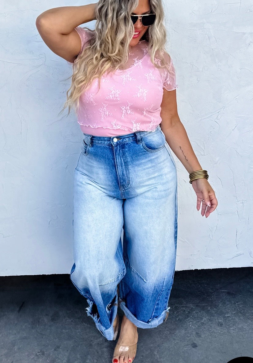 Woman wearing a pink lace top and blue jeans against a white wall.