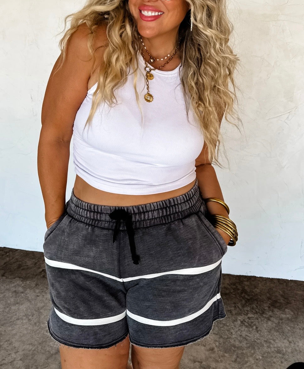 Woman wearing a white tank top and gray shorts with white stripes indoors.