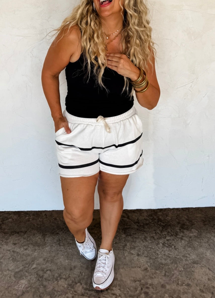 Woman wearing a black tank top and white shorts with black stripes against a white wall.