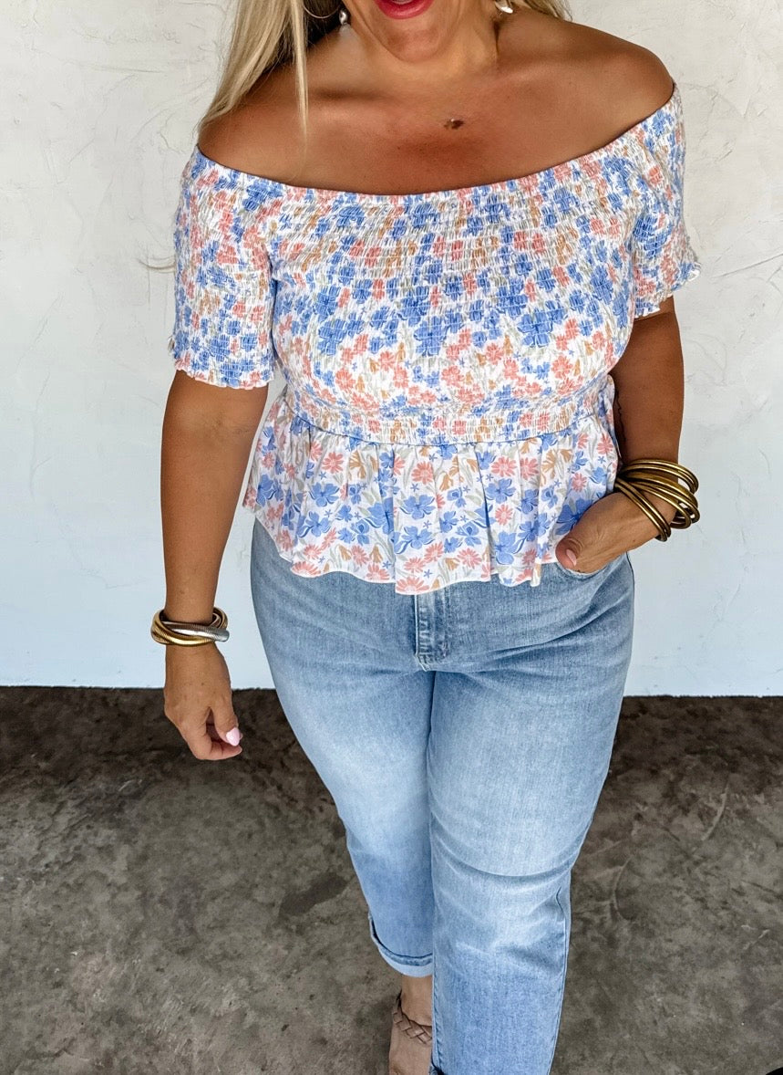 Woman wearing a floral off-shoulder top and blue jeans against a white wall.