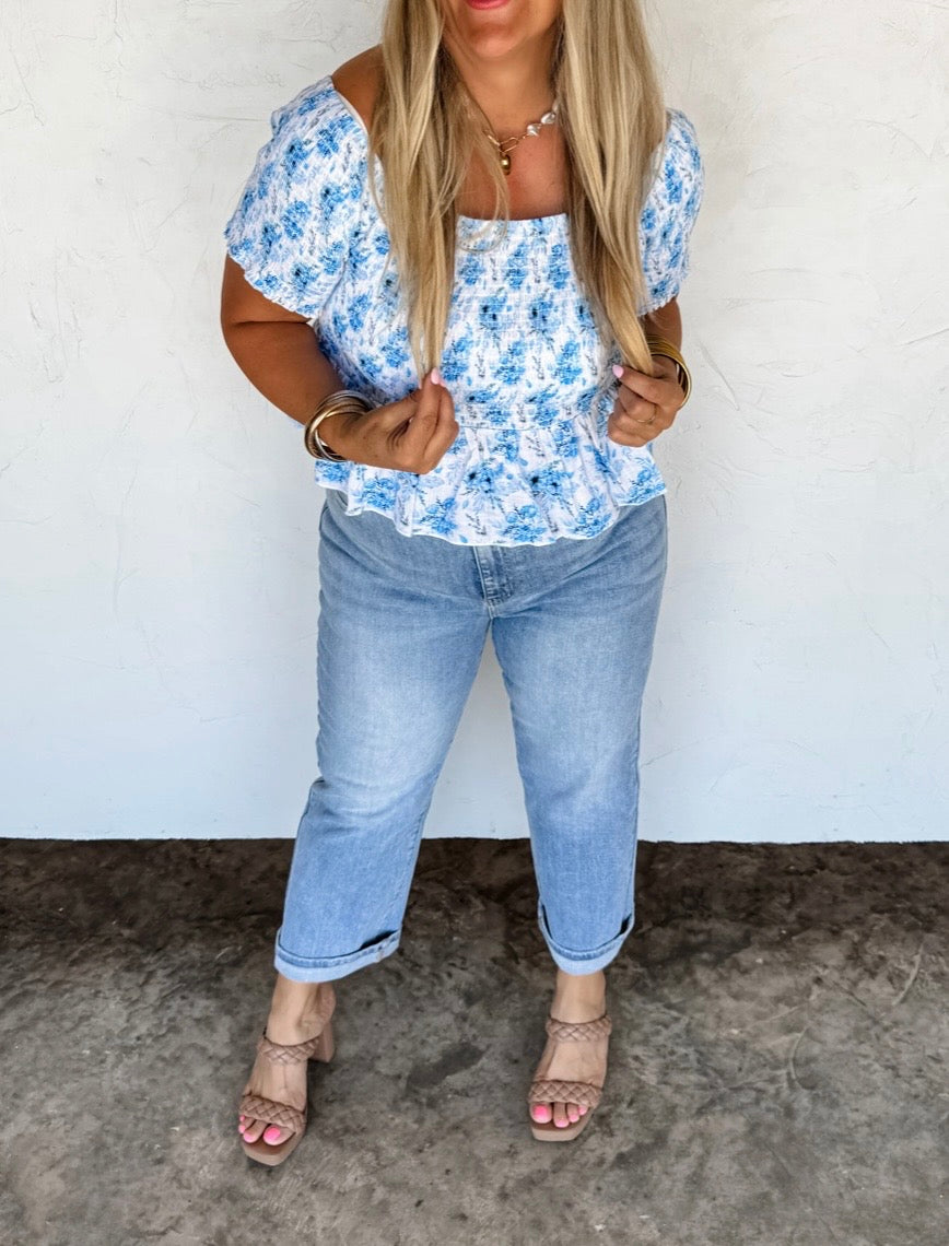 Woman wearing a blue floral top and jeans against a white wall.