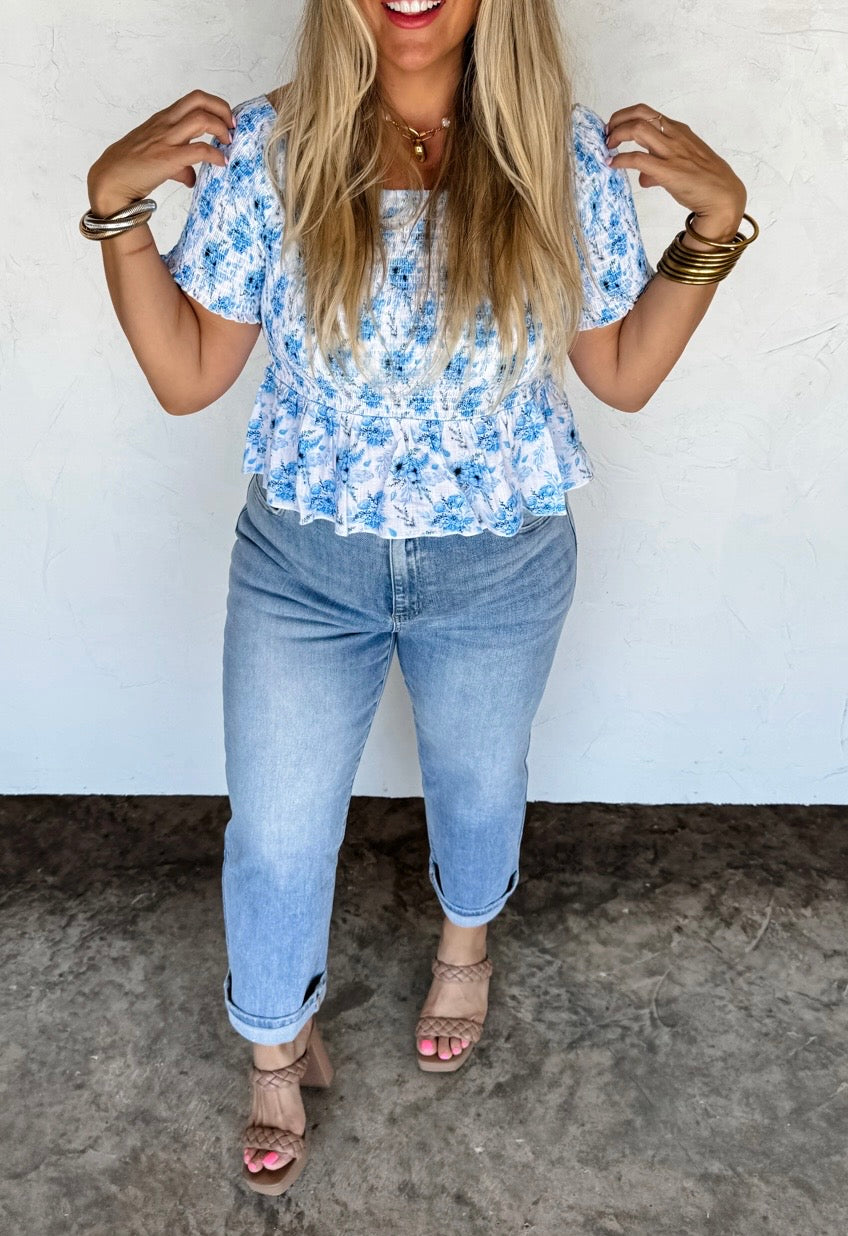 Woman wearing a blue floral top and jeans against a white wall.