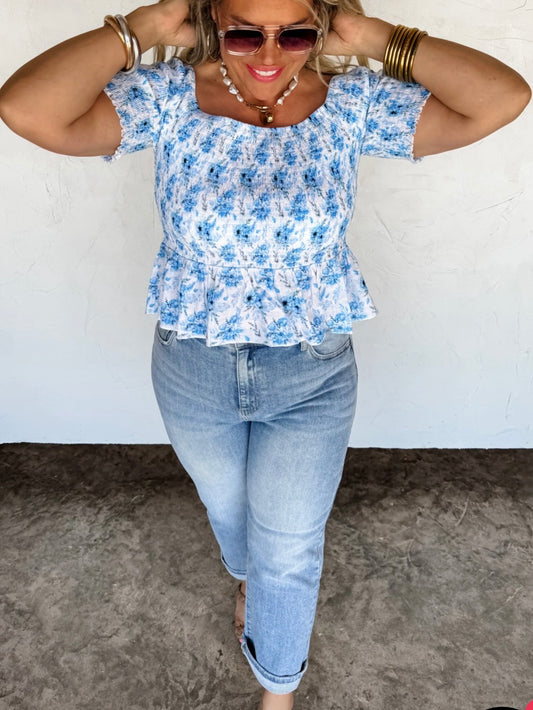 Woman wearing a blue floral top and jeans against a white wall.