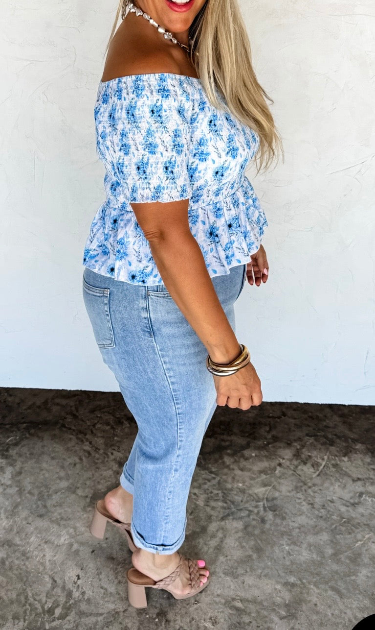 Woman wearing a blue floral off-shoulder top and jeans against a white wall.