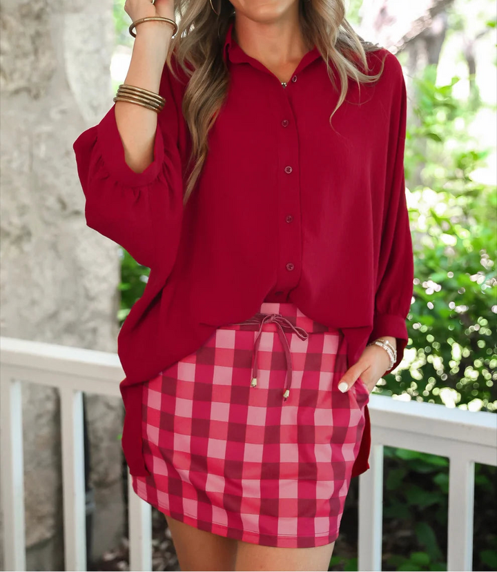 A woman posing outdoors wearing a long-sleeve red button-up shirt and a pink and cranberry gingham skort.