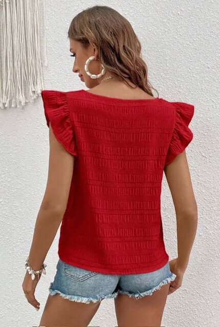 Woman wearing a red textured top with ruffled shoulders against a white wall.