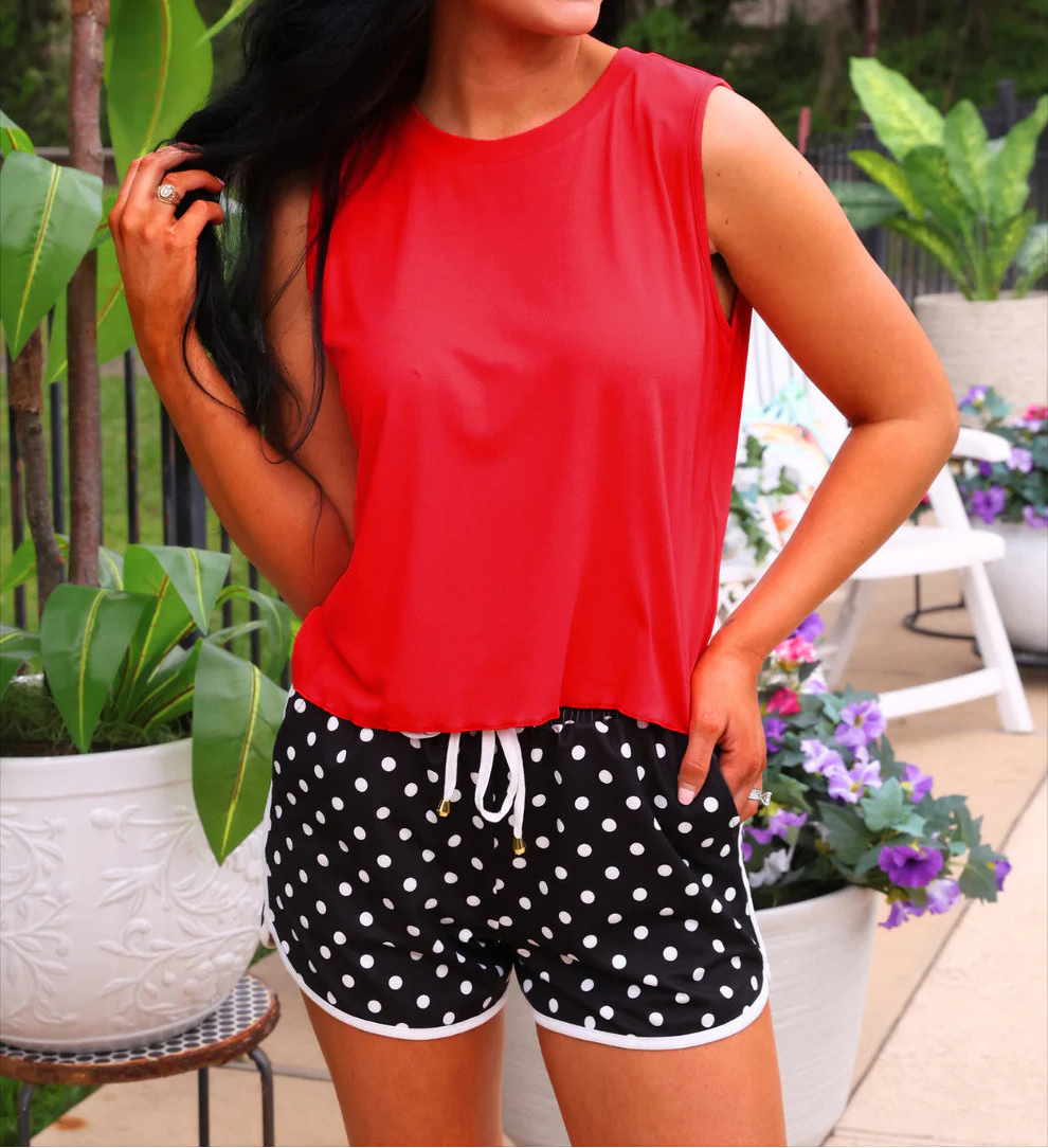 Woman wearing a red top and black polka dot shorts outdoors with plants and furniture in the background.