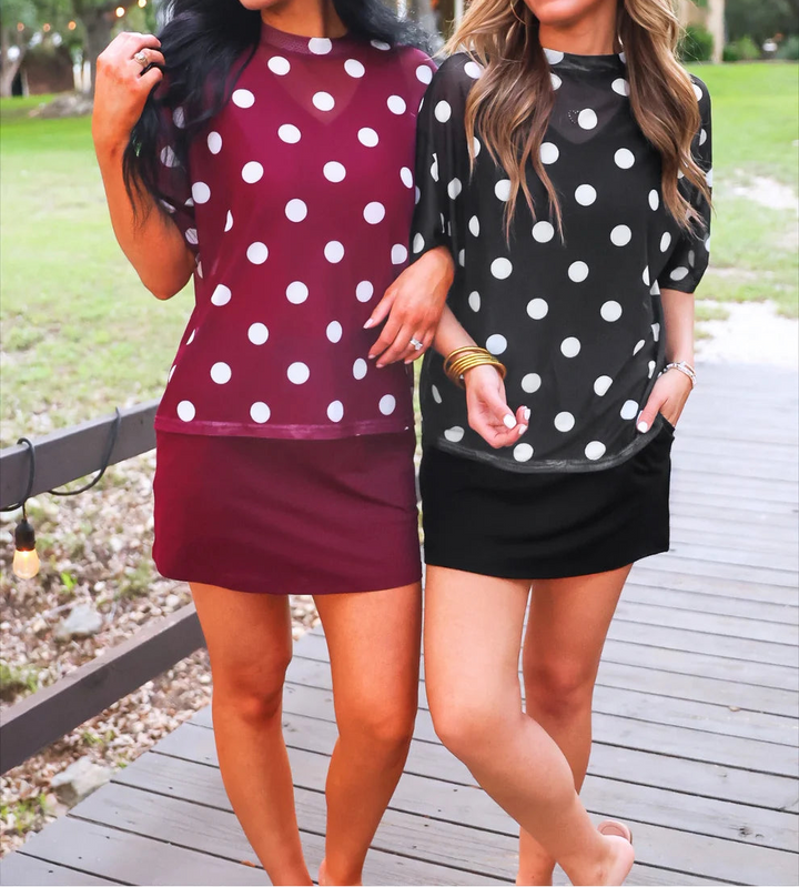 Two women wearing polka dot tops and skirts standing on a wooden deck with trees in the background.