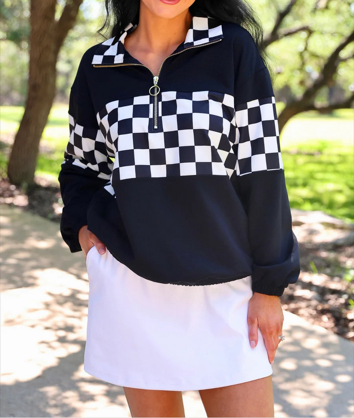 A woman wearing a black and white checkered quarter-zip sweatshirt and a white tennis skirt, standing outdoors on a sunny day with trees in the background.