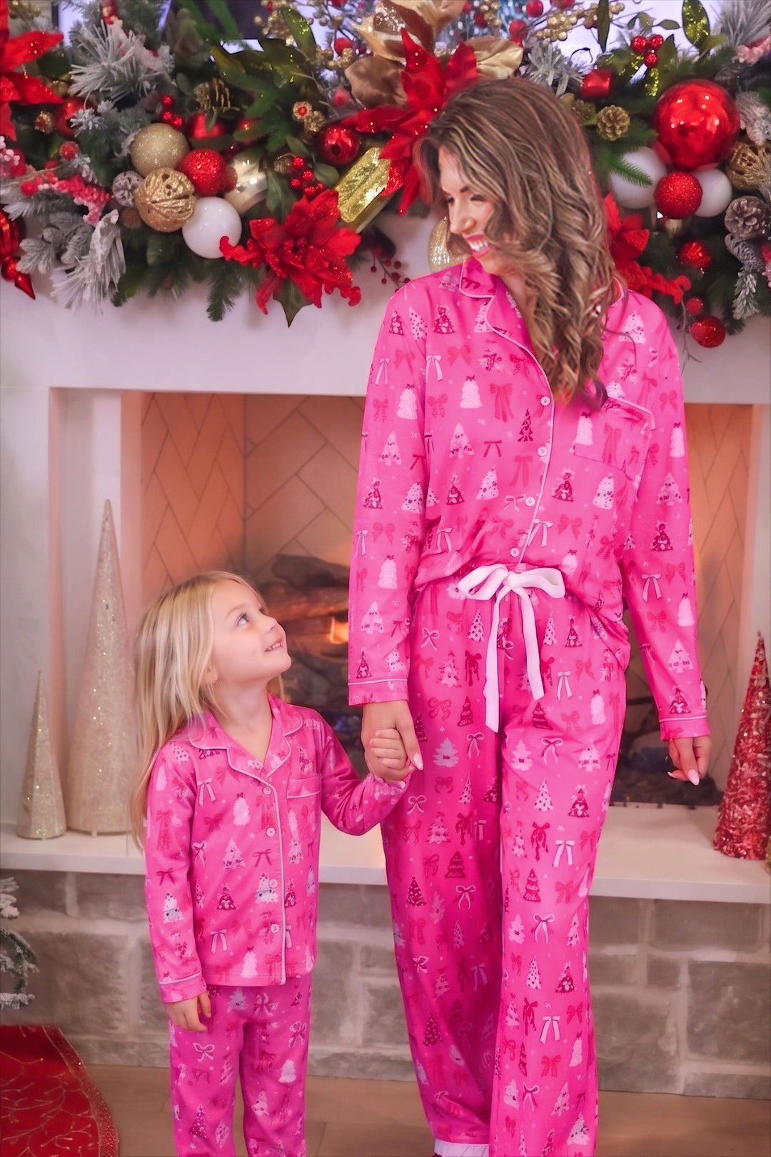 A woman and a young girl wearing matching bright pink Christmas-themed pajamas with a white bow tie, standing in front of a decorated fireplace.