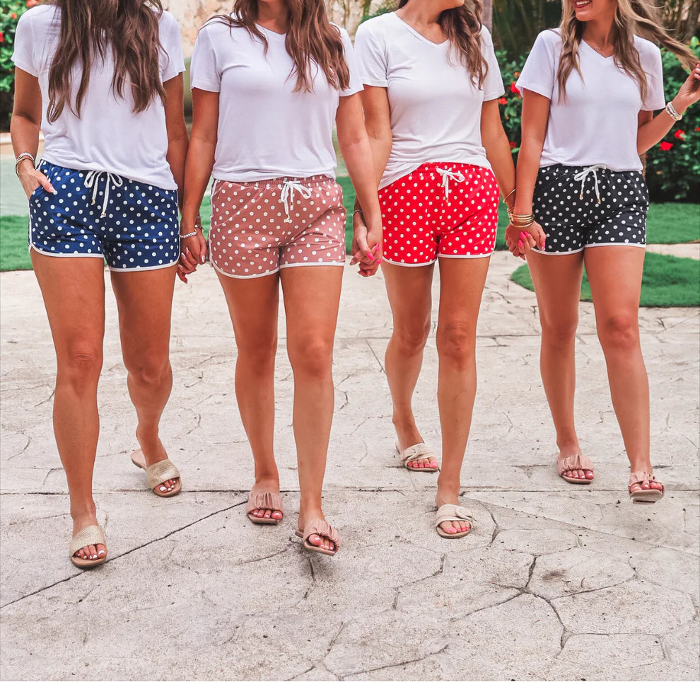 Four women standing together in front of palm trees wearing matching white t-shirts and colorful shorts.