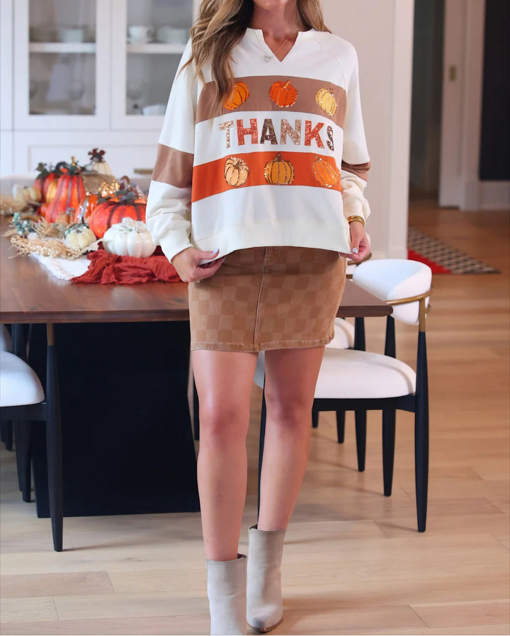 A woman stands in a dining room wearing a white long-sleeve V-neck sweater with a brown and orange horizontal stripe pattern across the chest featuring the word &