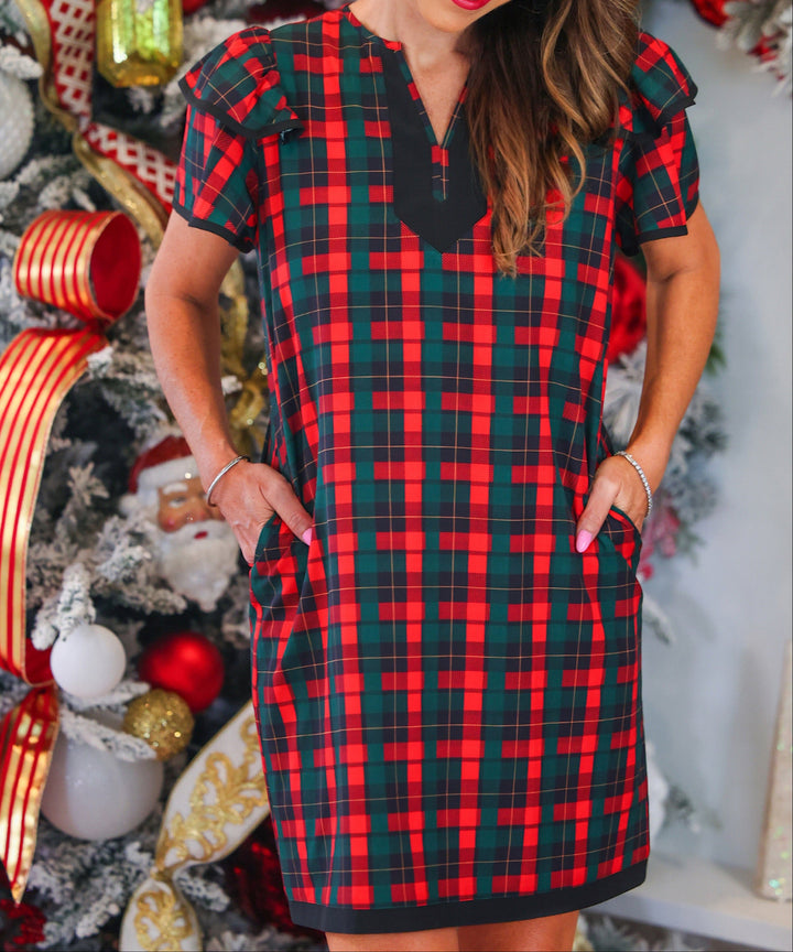A woman wearing a red and green plaid short-sleeve holiday dress with a black V-neckline, standing in front of a decorated Christmas tree.