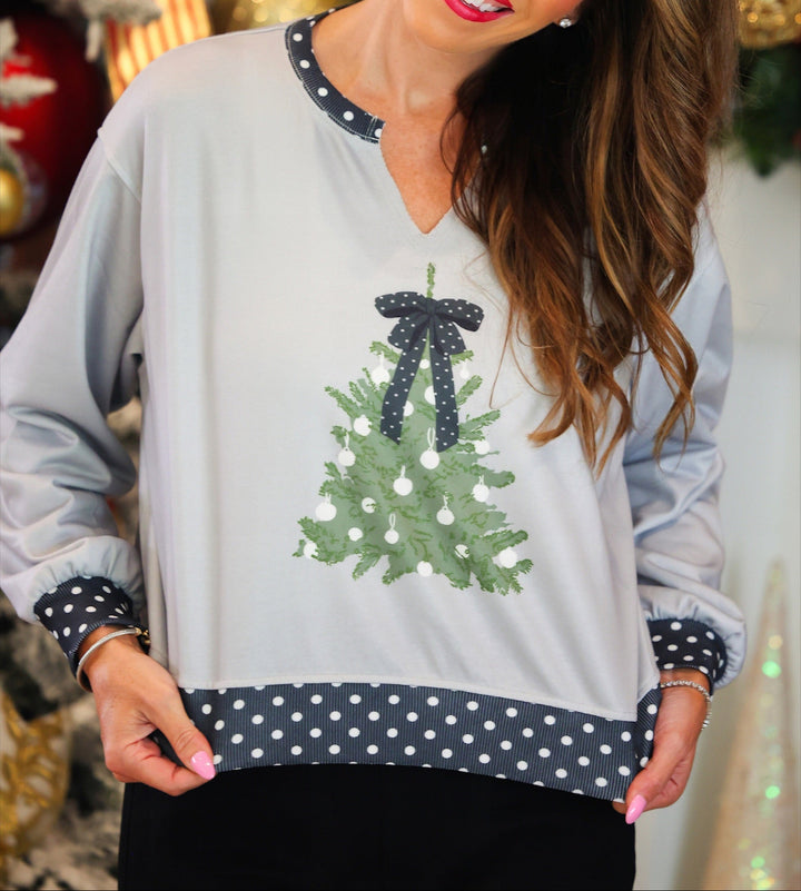 A woman wearing a light gray long-sleeve sweater with a green Christmas tree graphic and navy and white polka dot trim, posed in front of a festive holiday background.