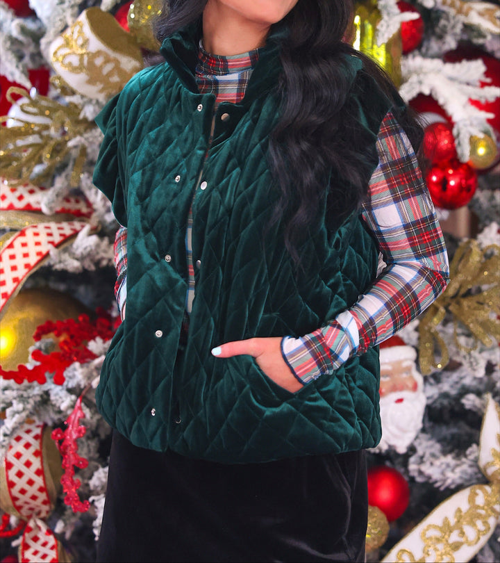 A woman wearing a dark green velvet quilted vest over a red, green, and white plaid long-sleeve shirt, standing in front of a decorated Christmas tree.