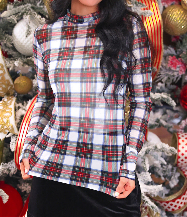 A woman wearing a long-sleeve plaid mock neck top with a white base and red, green, and black tartan pattern, posed in front of a decorated Christmas tree.