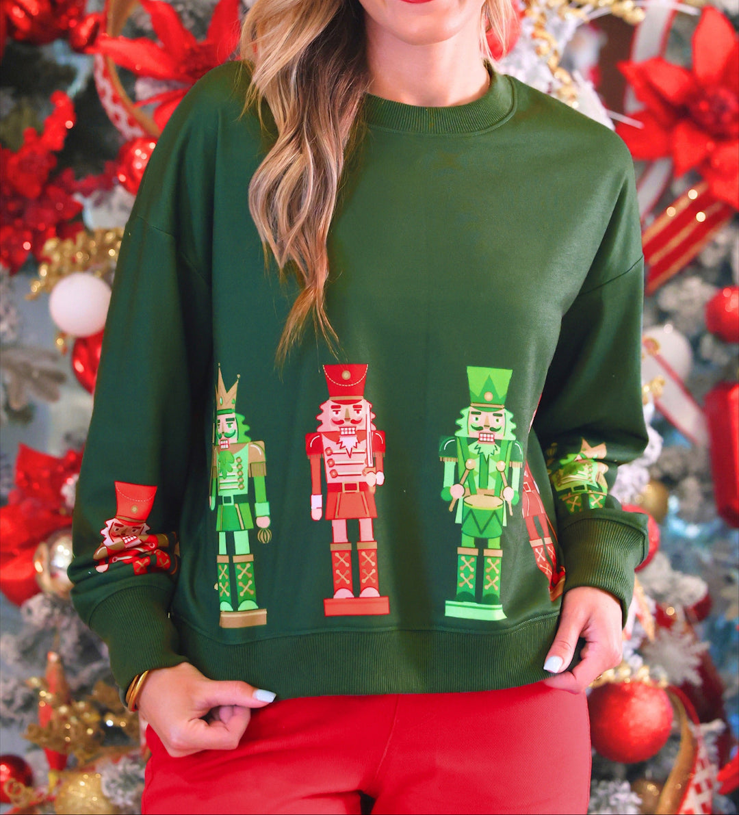 A woman wearing a dark green crewneck sweatshirt with a graphic of three nutcracker figures, posed in front of a decorated Christmas tree.