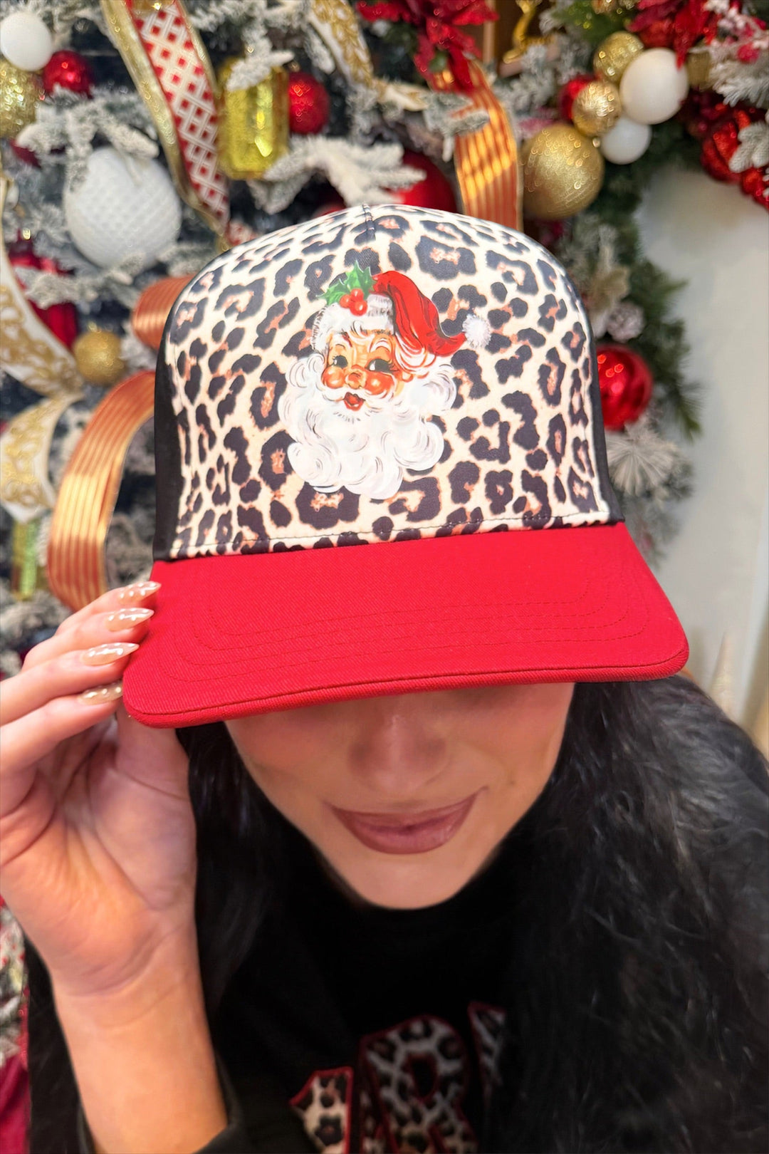 A person wearing a red and leopard-print baseball cap with a Santa Claus graphic, posed in front of a decorated Christmas tree.