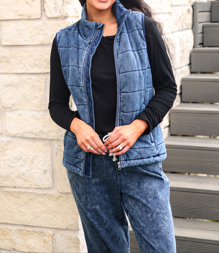 A woman wearing a blue quilted zip-up vest over a black long-sleeve top and matching blue pants, standing outdoors against a light stone wall and dark wooden steps.