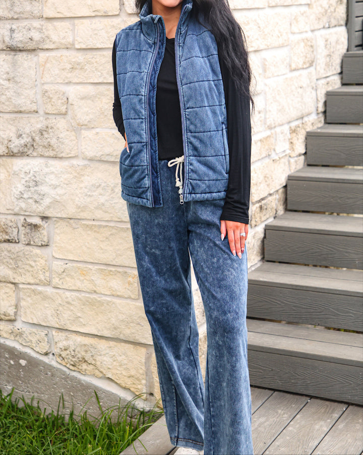 A woman wearing a blue quilted zip-front vest over a black long-sleeve top and matching blue wide-leg pants, standing outdoors against a light stone wall with gray steps beside her.