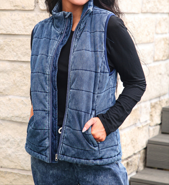 A woman wearing a blue quilted zip-up vest over a black long-sleeve top, standing against a light stone wall.
