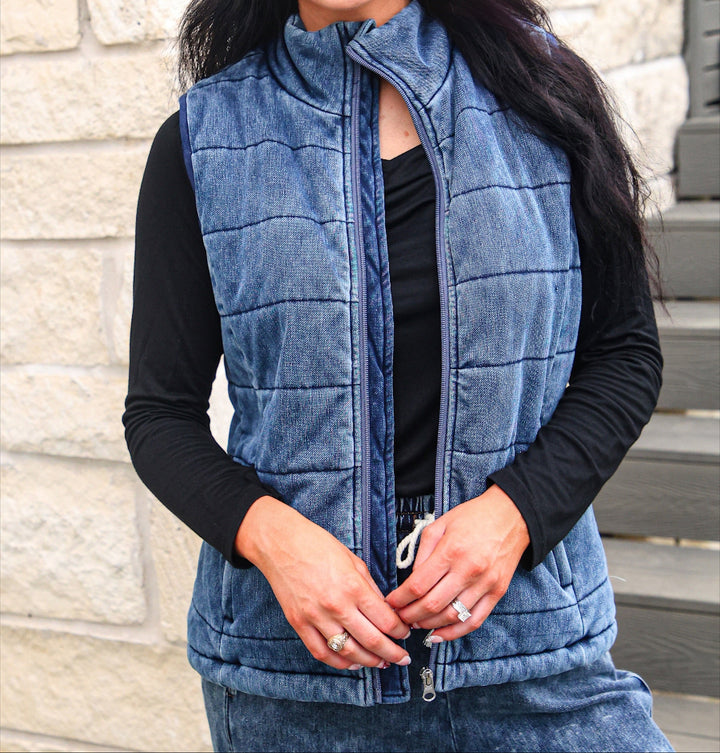 A woman wearing a blue quilted zip-front vest over a black long-sleeve top, paired with a white baseball cap covered in colorful rhinestones that spell &