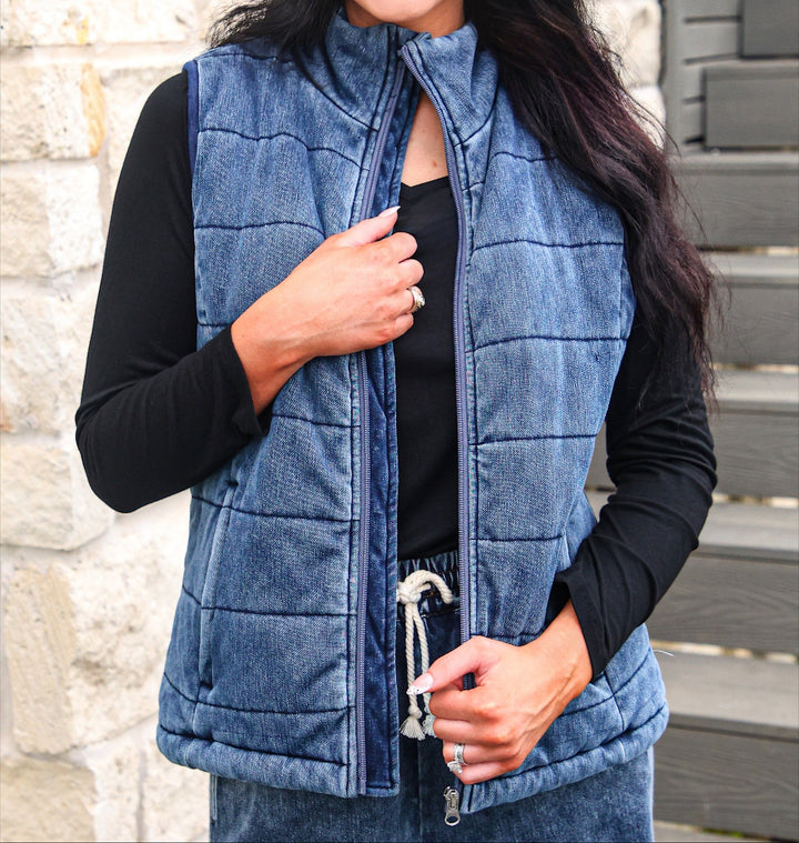 A smiling woman wearing a blue quilted zip-front vest over a black long-sleeve top, with a colorful polka-dot cap that says &