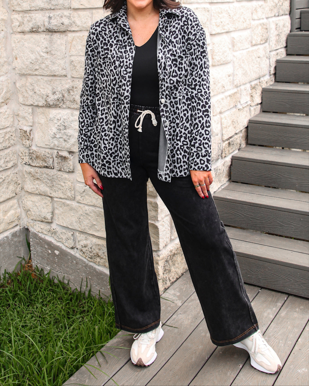 A woman stands outdoors on a wooden deck in front of a light stone wall and gray steps, wearing a black-and-white leopard print button-up shirt over a black top and black wide-leg pants with white sneakers.