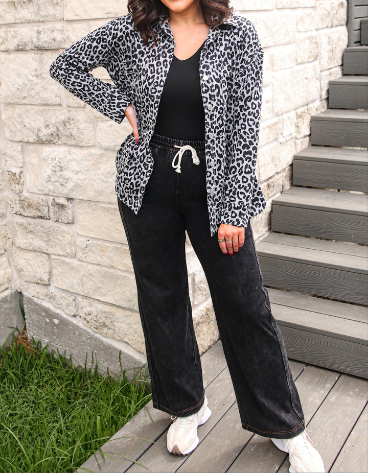 A woman stands outdoors on a wooden deck beside a light stone wall and gray steps, wearing a black-and-white leopard print button-up shirt over a black top, black wide-leg pants with a white drawstring, and white sneakers.