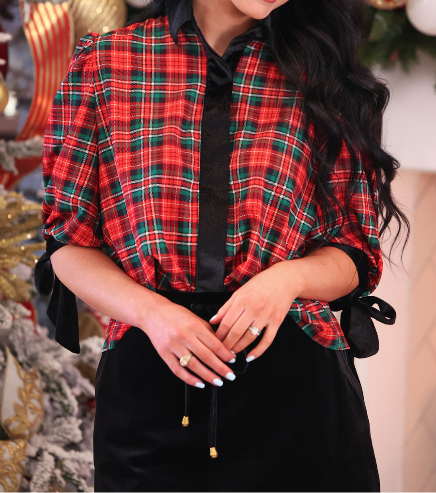 A woman wearing a festive red, green, and black plaid button-up shirt with a black satin tie-neck and black bow-tie cuffs, paired with a black skort, posed in front of a decorated Christmas tree.