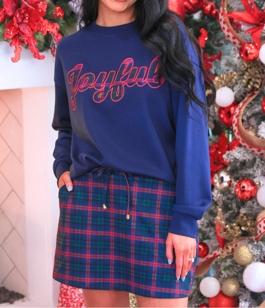 A woman stands in front of a decorated Christmas tree wearing a navy blue crewneck sweatshirt with &