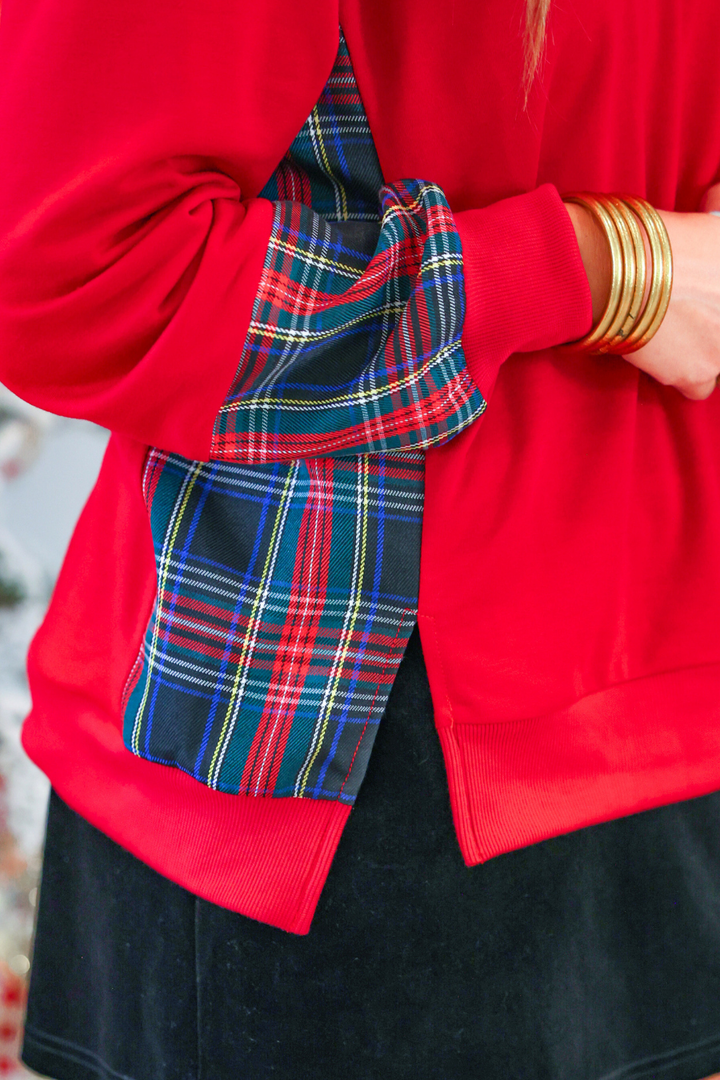 A close-up of a person wearing a bright red sweatshirt with a large plaid tartan bow tied around the back or side, paired with a black skirt and gold bangles.