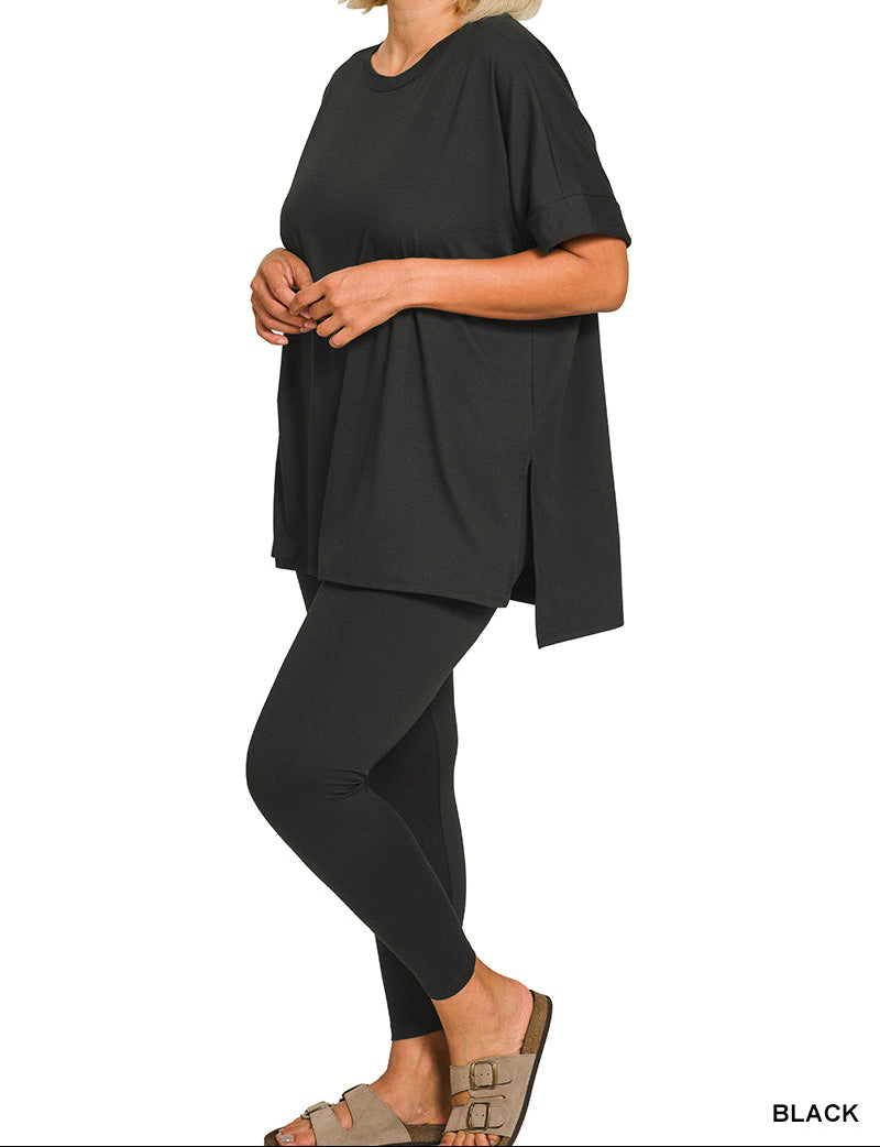 A woman modeling a black oversized short-sleeve top with matching black leggings and tan slide sandals, shown against a plain white background. The word &
