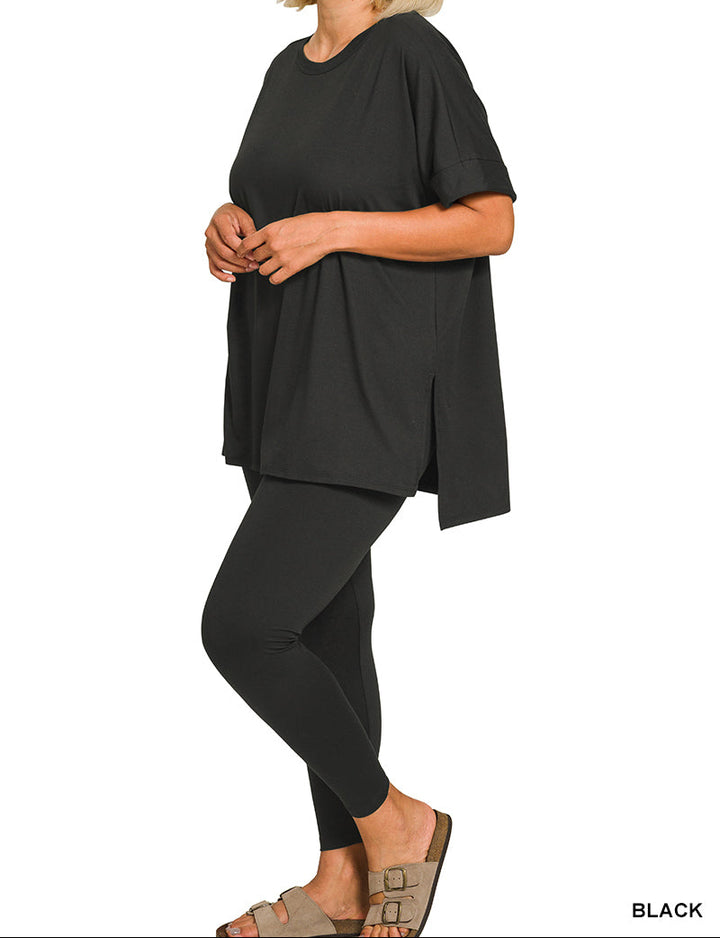 A woman modeling a black oversized short-sleeve top with matching black leggings and tan slide sandals, shown against a plain white background. The word &