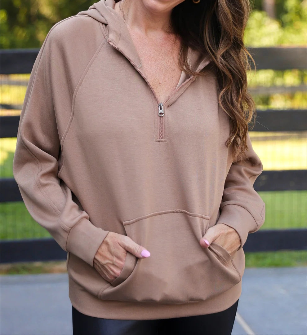 Woman wearing a mocha hoodie with a zipper, standing outdoors with greenery in the background.