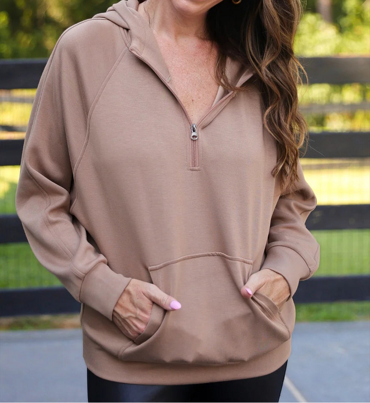 Woman wearing a mocha hoodie with a zipper, standing outdoors with greenery in the background.