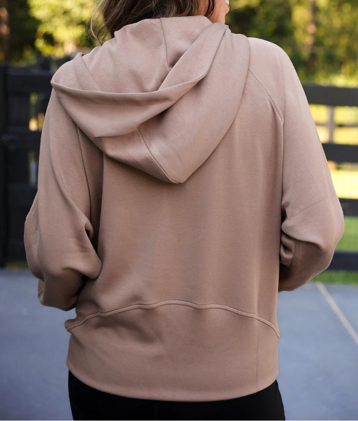 A woman viewed from behind wearing a mocha brown pullover hoodie with a relaxed fit, set against an outdoor background of trees and a fence.