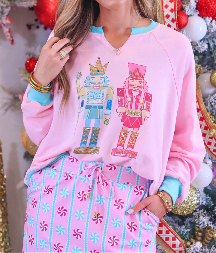 A woman wearing a pink crewneck sweatshirt with a nutcracker graphic and matching candy-print pajama pants, posed in front of a decorated Christmas tree.