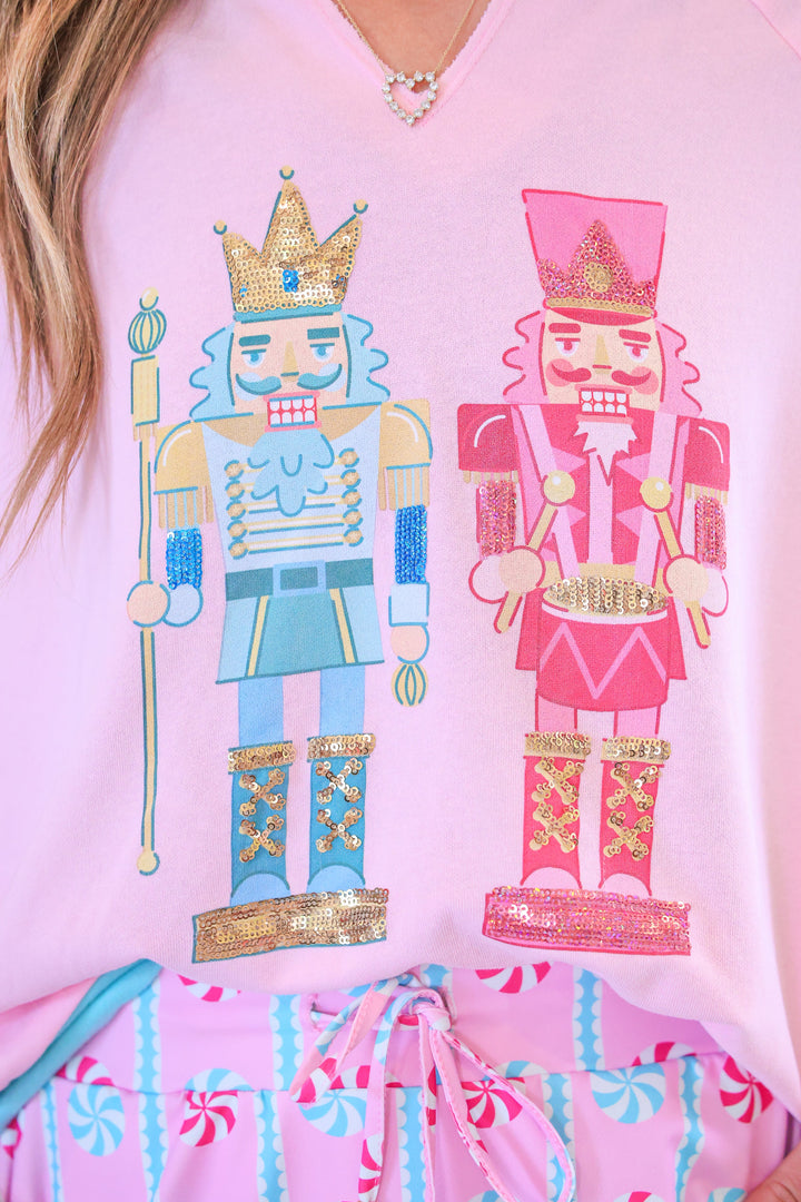 A close-up of a light pink V-neck t-shirt featuring a graphic print of two nutcracker figures, one in light blue with gold accents and one in pink with gold accents. The shirt is shown on a model wearing a pink patterned skirt.