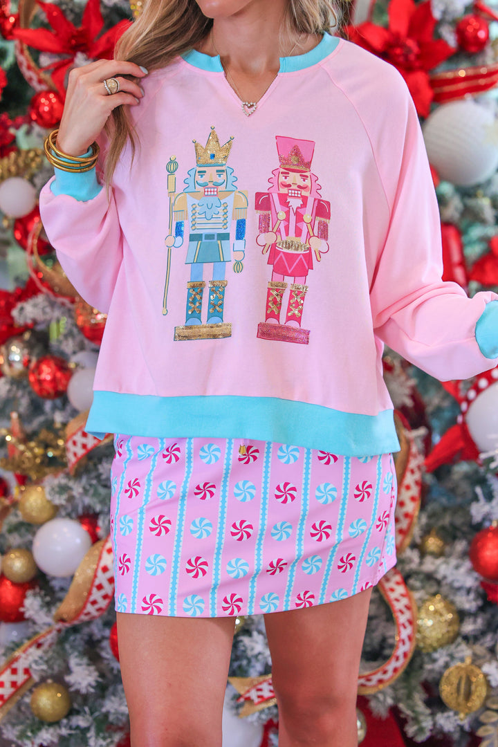 A woman wearing a pink crewneck sweatshirt with a nutcracker graphic and a pink candy cane patterned skirt, posed in front of a decorated Christmas tree.