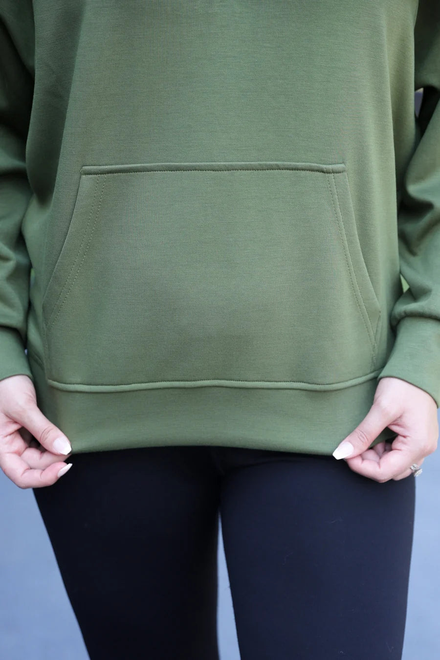 A close-up of a person wearing an olive green pullover hoodie with a front kangaroo pocket, paired with black leggings.