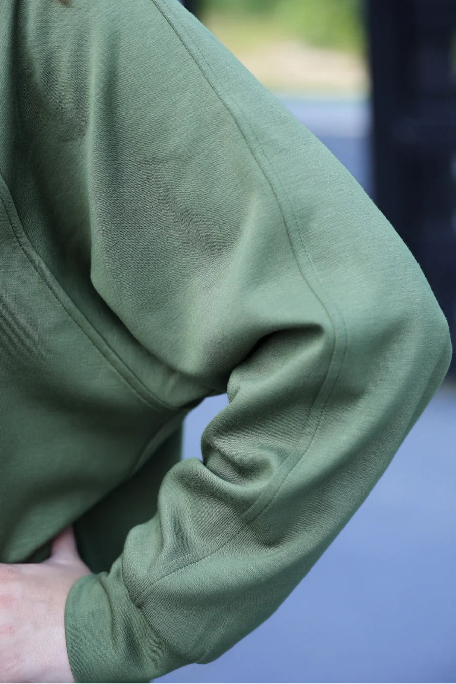 Close-up of a person wearing an olive green long-sleeve top, showing the fabric texture and sleeve detail against a blurred outdoor background.