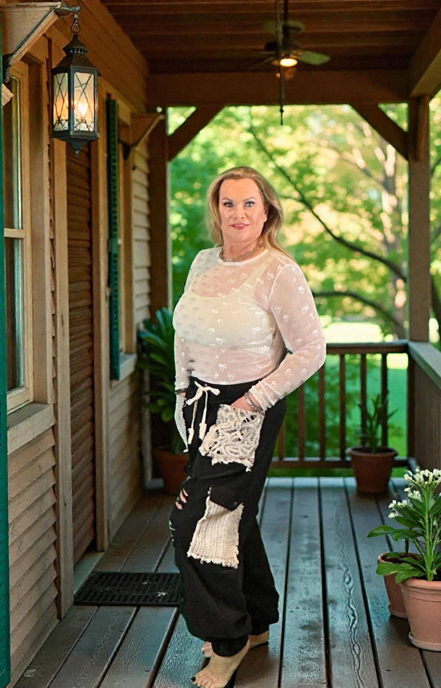 Woman standing on a wooden porch with plants and a green landscape.