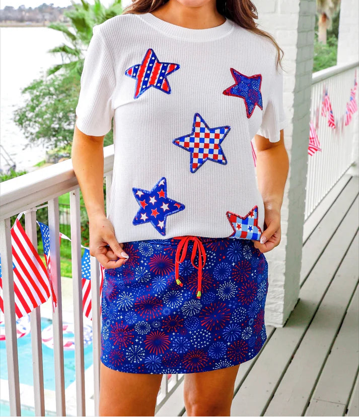 Woman wearing a white t-shirt with red, white, and blue star patterns and a blue skirt with red accents on a porch.